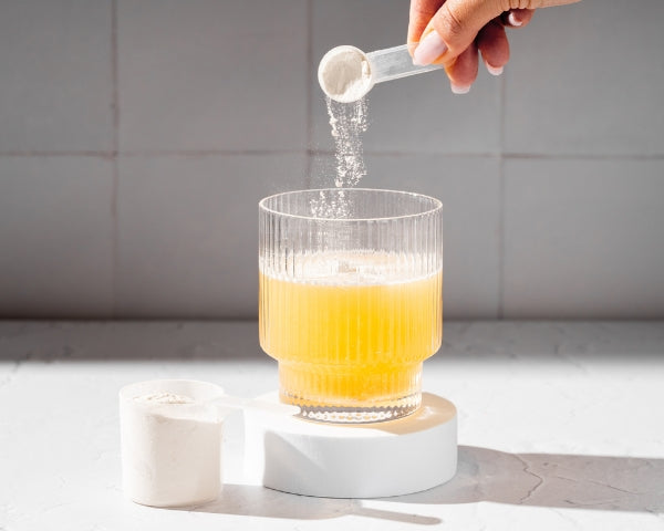 Kollagen Pulver wird mit Messlöffel in ein Glas mit orangem Getränk gegeben zur Unterstützung von Haut Haaren und Gelenken